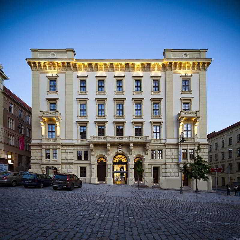 Grand Palace Brno Luaran gambar The photo shows a historic building with a classical architectural style. It features a cream-colored façade with decorative elements, large windows, and ornate detailing around the entrance. The building is four stories high and is illuminated in th